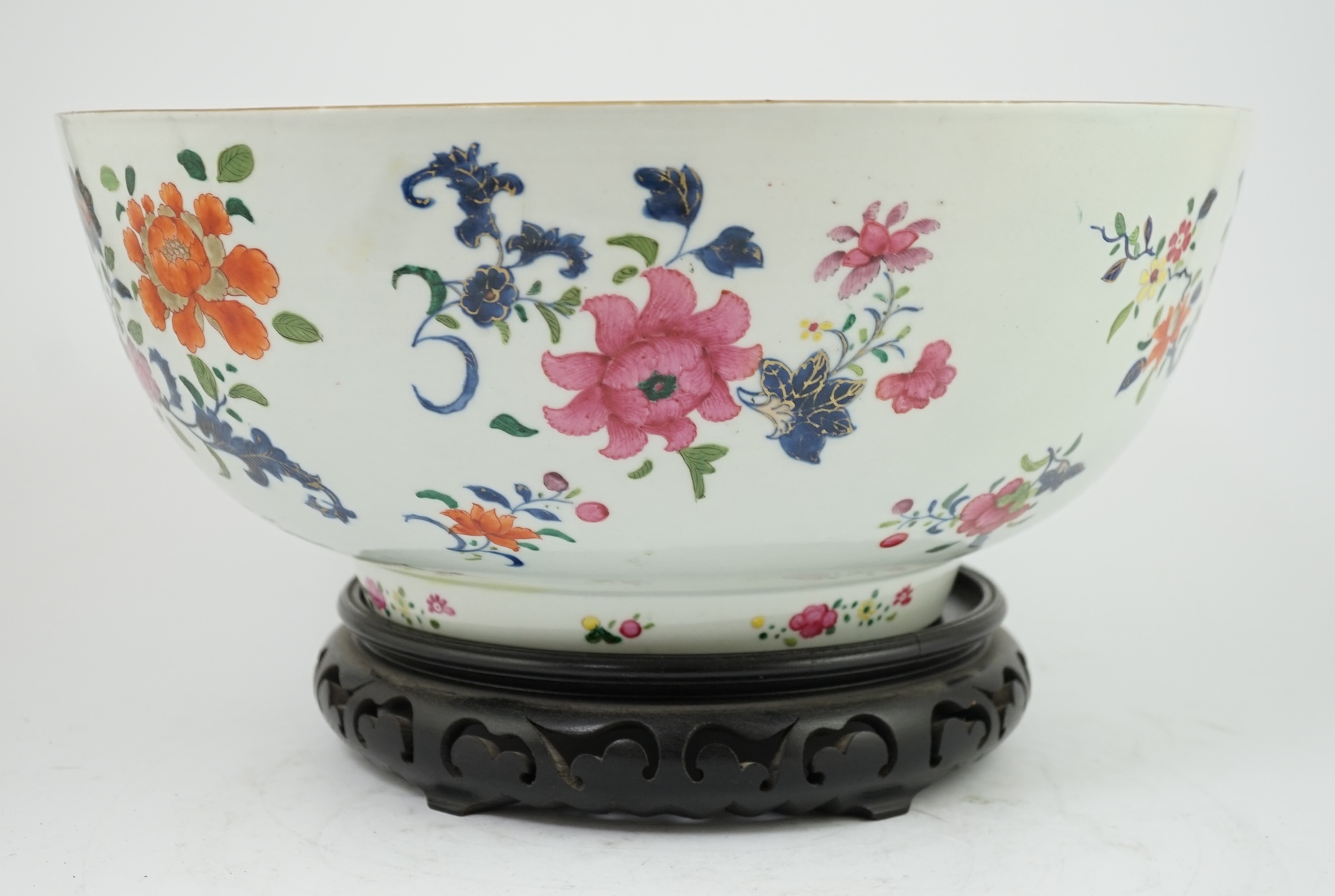 A large Chinese famille rose punch bowl, Qianlong period, the exterior painted with flowers in a fenced garden, the interior with a flower bouquet within a garland of flowers, 40.5cm diameter, wood stand. Condition - som